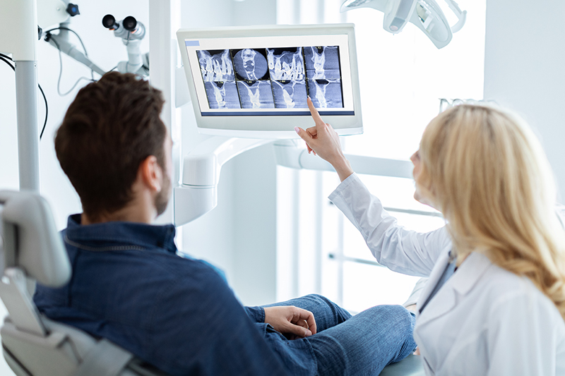 Modern dental clinic concept. Blonde woman dentist having conversation with patient in dental chair, poitning at LED display, showing results of treatment or explaining procedure